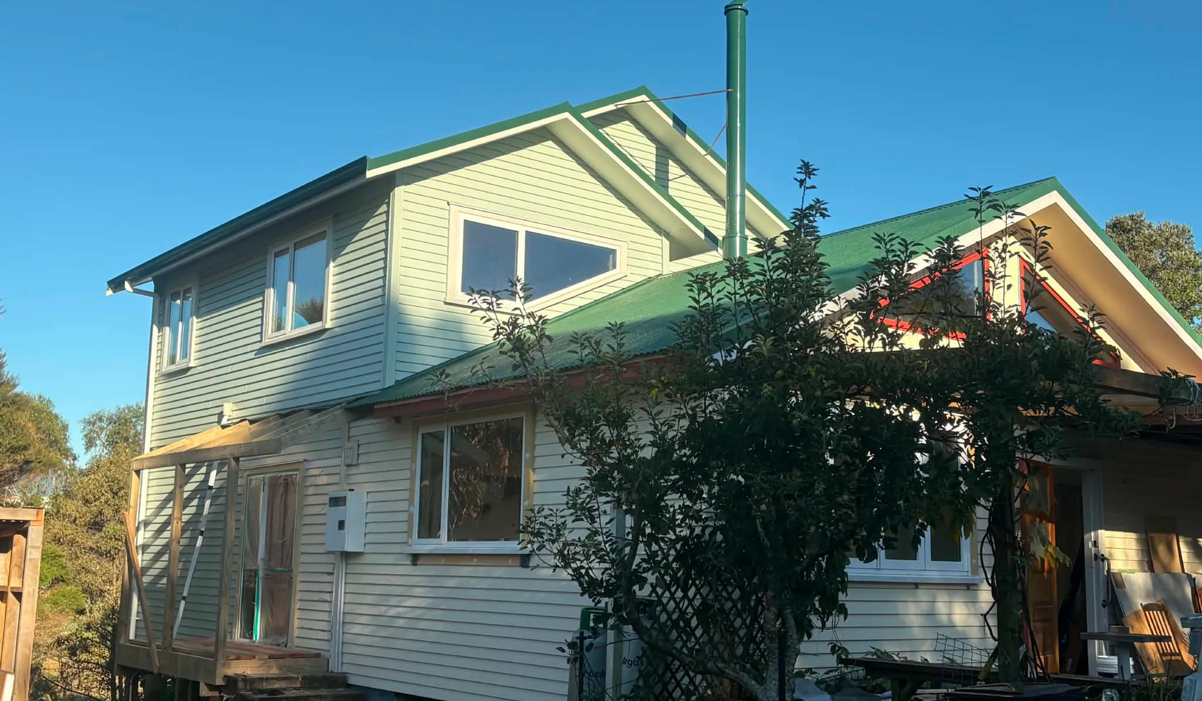 Mid-build view of the Government Road two-storey extension — new wing in white weatherboard, original red-roof house at right, late afternoon