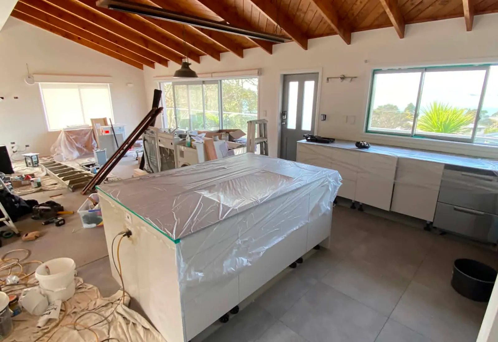 Late-stage finishing — new kitchen island wrapped for protection, cabinets installed, dropsheets covering the new floor