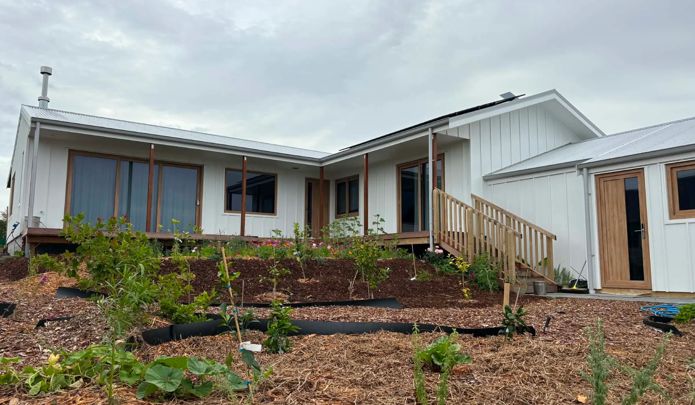 Finished new build at 73 Rangitahi — single-storey, white vertical cladding, deck and steps, with mulched gardens out front.