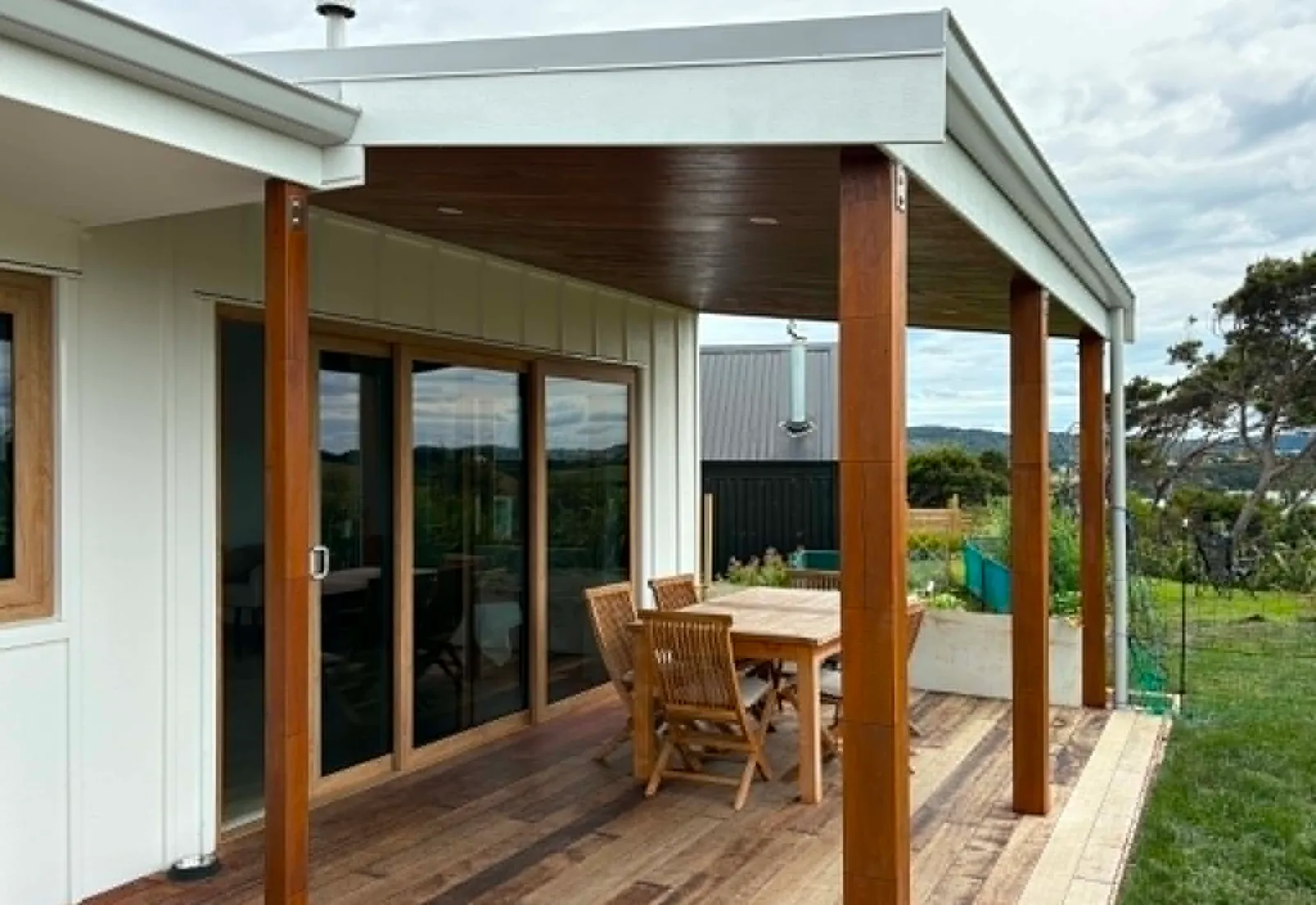 Finished covered hardwood deck with a dining setting and chunky timber posts, attached to a white weatherboard Raglan home with hill views beyond