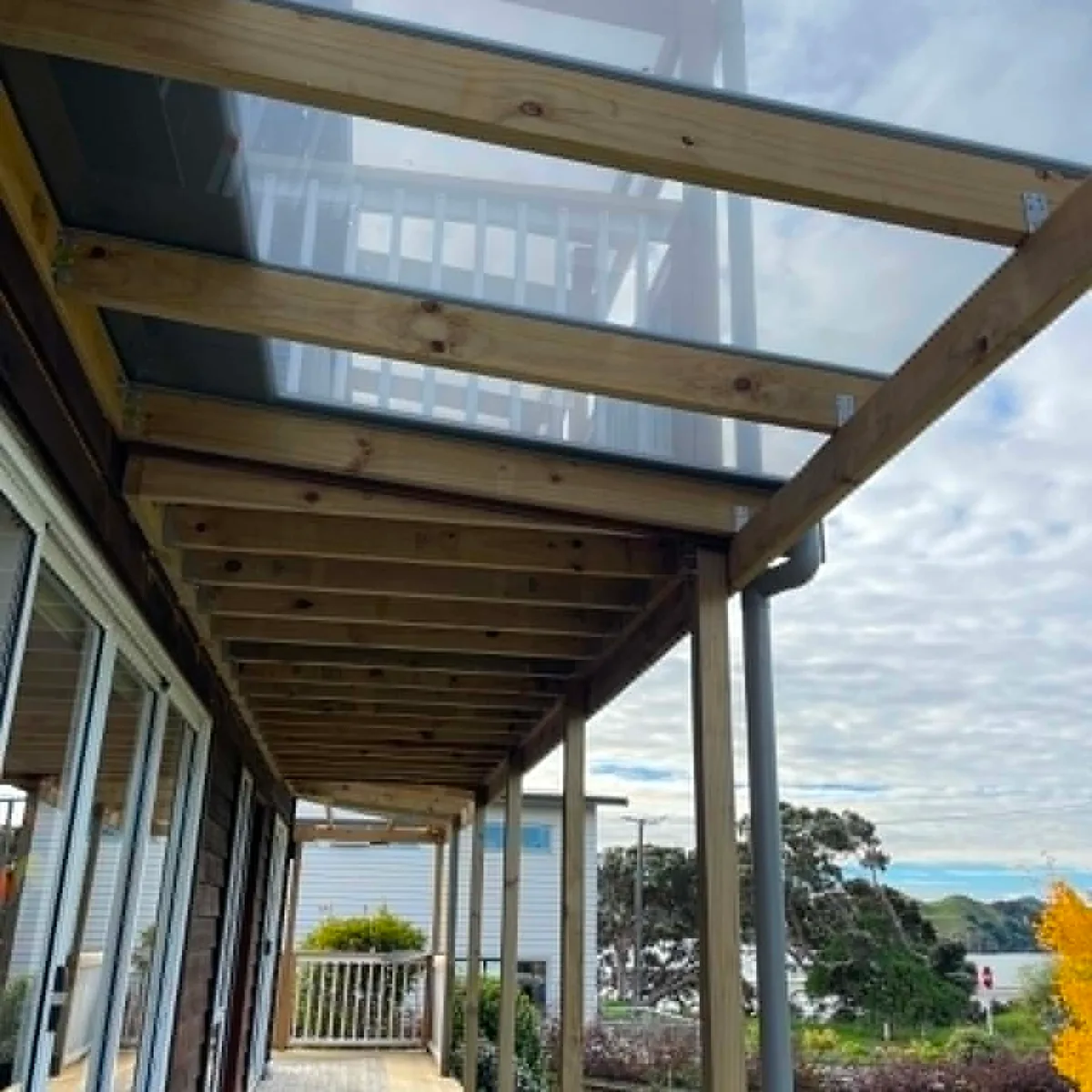Covered walkway along the side of a Raglan home, with timber framing and a clear roof, harbour and hills visible beyond
