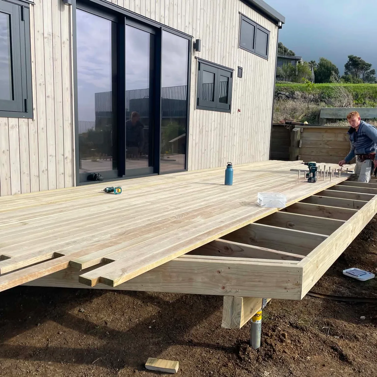 Mid-build deck construction outside a white weatherboard Raglan home, with joists installed on steel piles and decking boards going down