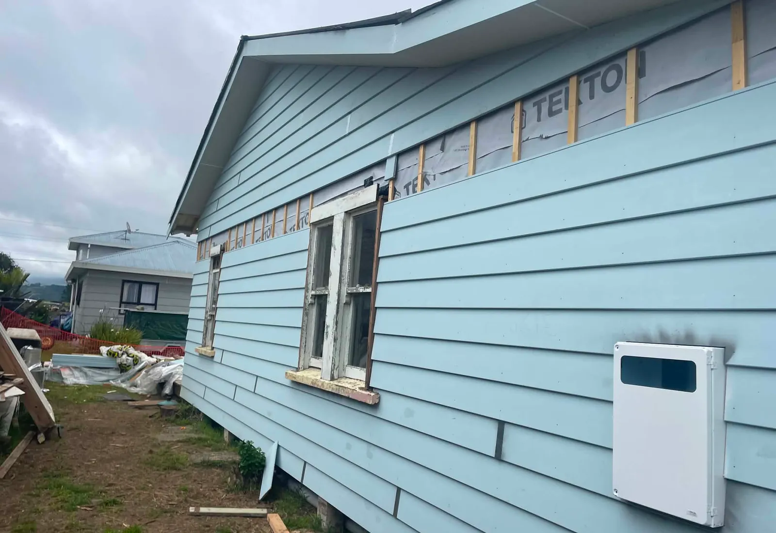 Pale blue weatherboard mid-recladding, with new boards installed below the eaves and building wrap still exposed above