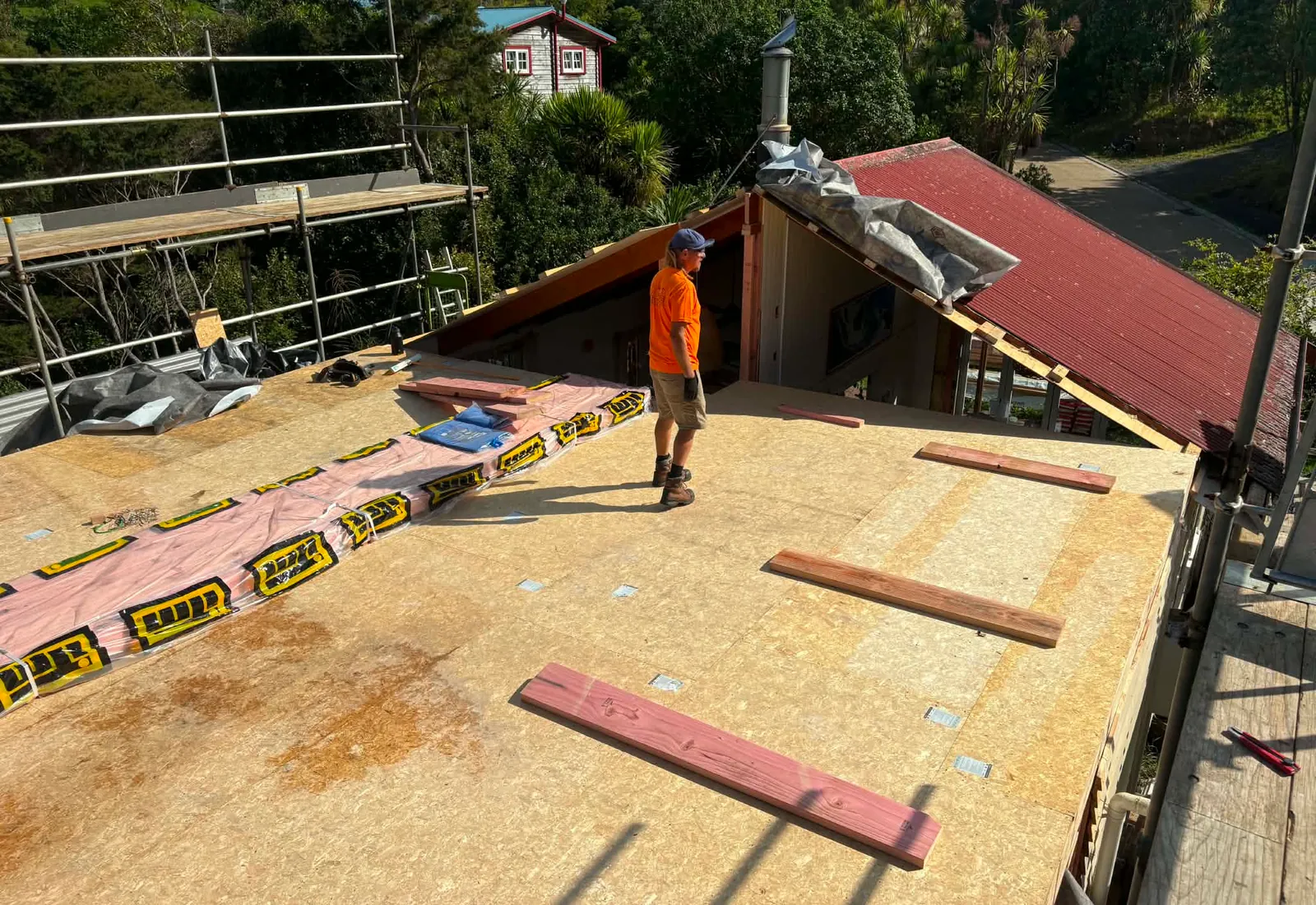 Roof-deck stage of a Raglan renovation — new plywood sheathing installed across the upper-level floor with the original red roof visible below