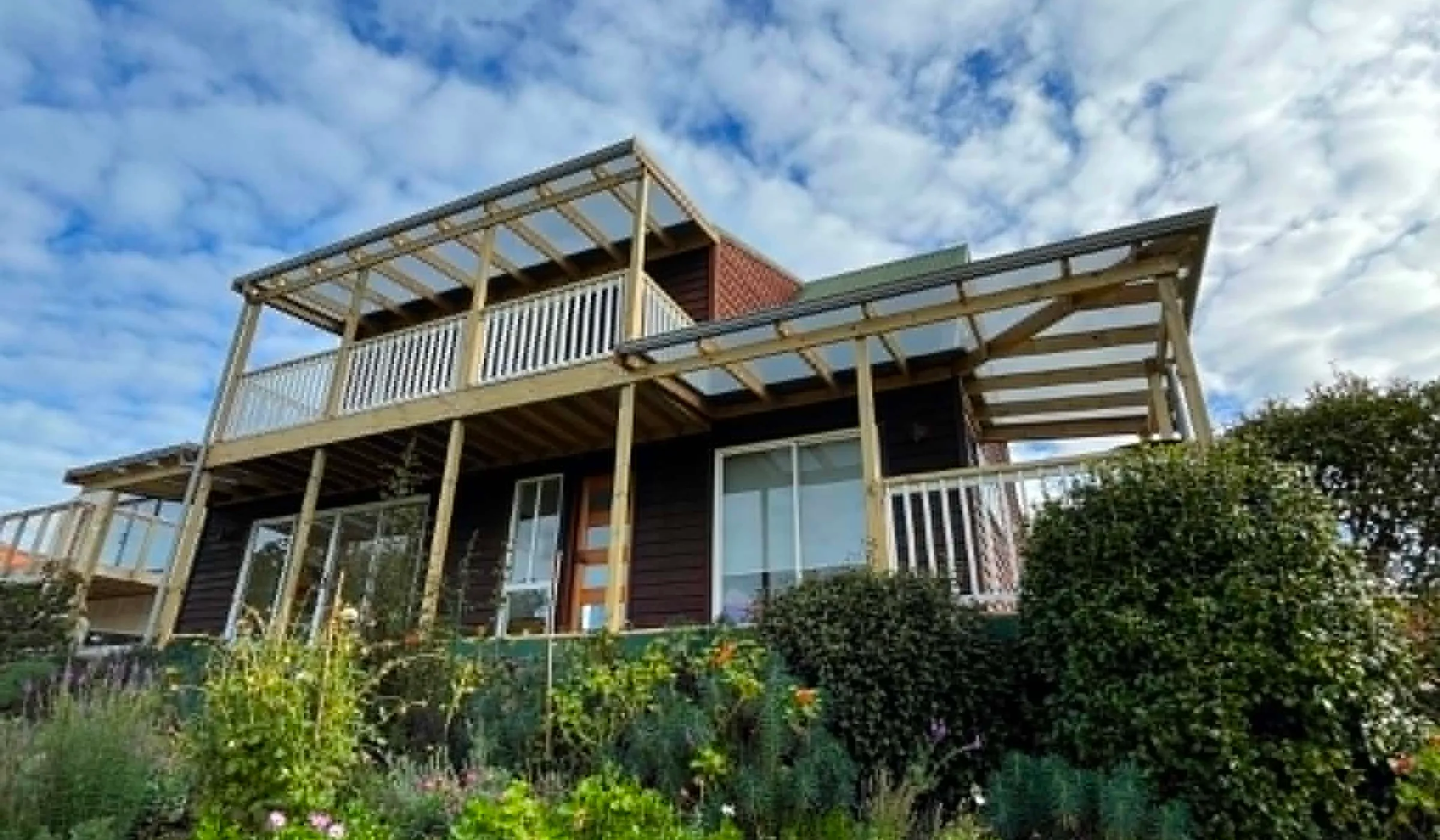 Renovated two-storey Raglan home with a timber upper-level balcony, dark ground-floor cladding, and a brick chimney rising through the centre