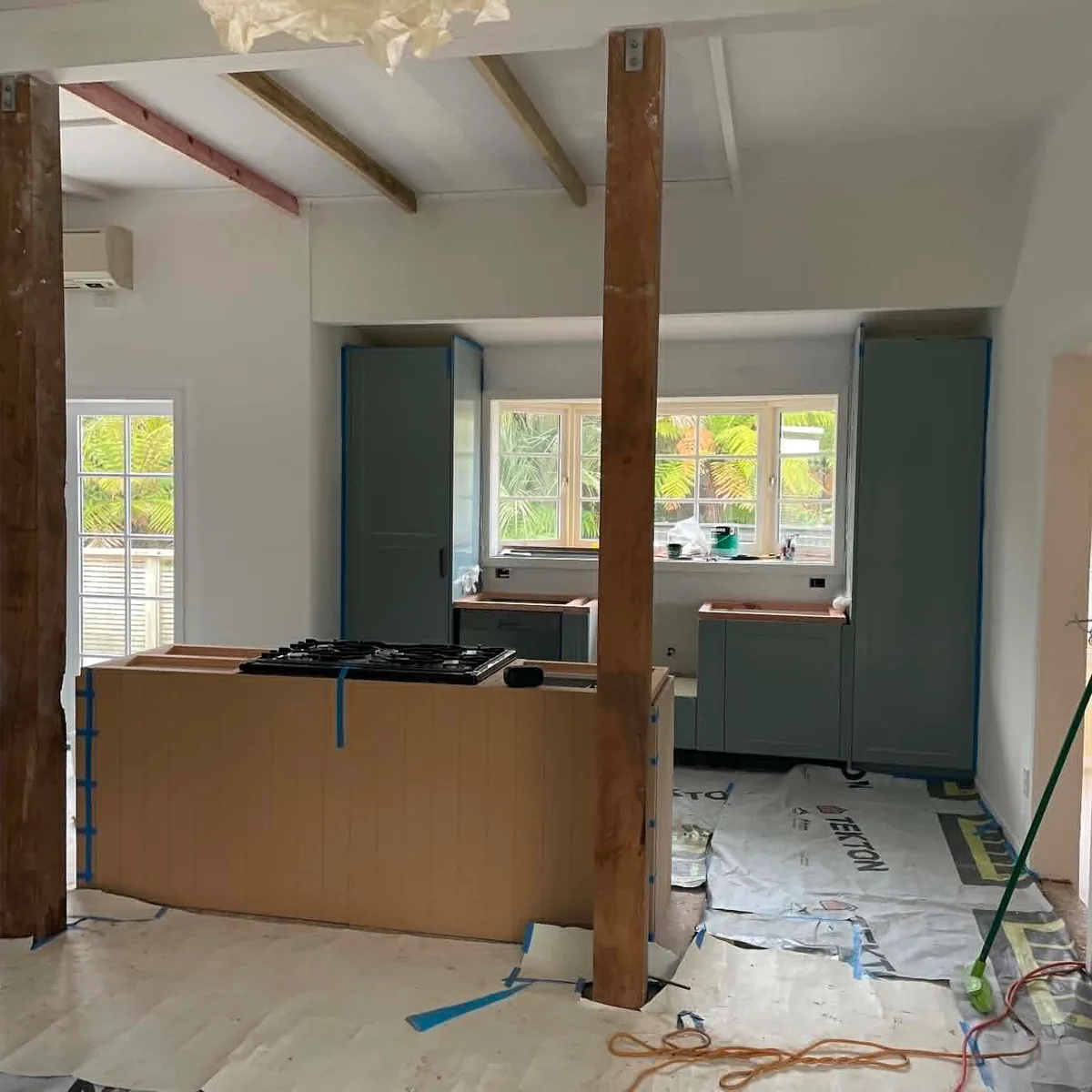 Kitchen renovation in the late stages — sage green cabinetry installed, cooktop in place, protective wrap on the floor